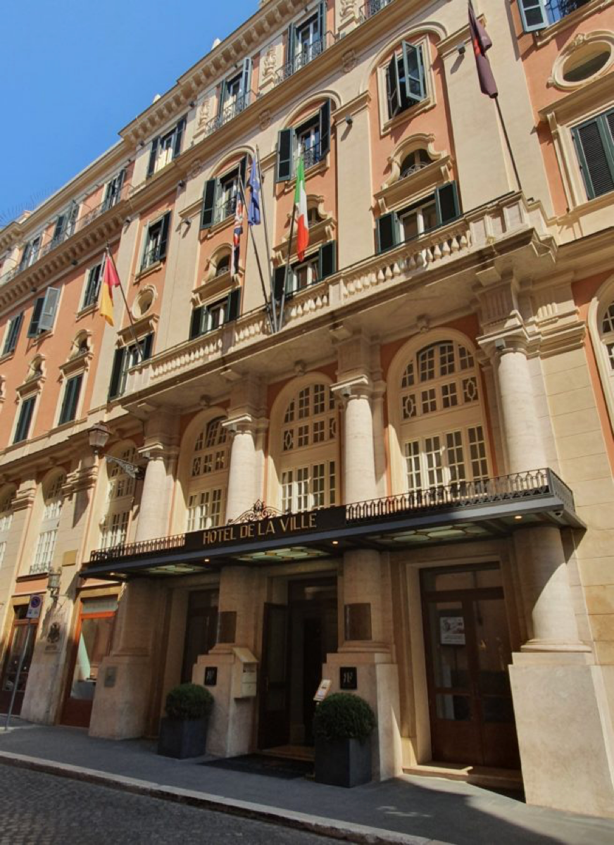 The entrance to the Hotel de la Ville in Rome, a Rocco Forte hotel with flags and a Renaissance style facade of salmon coloured and tan stone